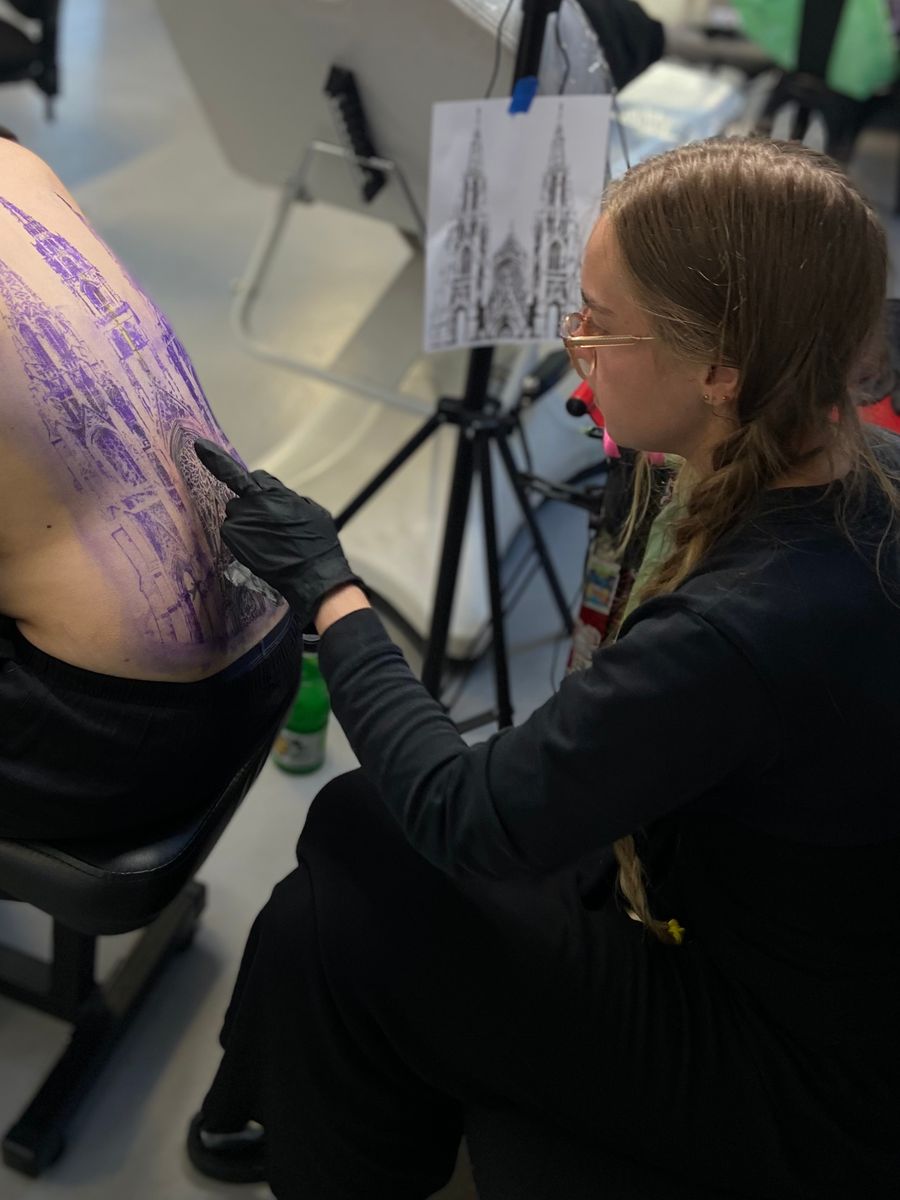 Pixi at work in the studio, gloved hand steadying a client's back as she checks a cathedral stencil against the reference drawing.