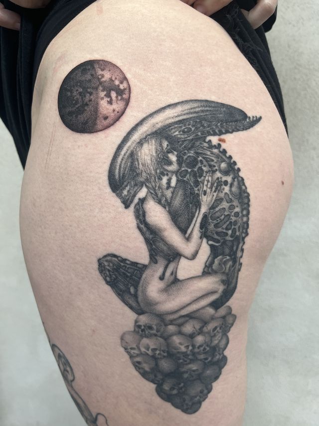 Black and grey thigh piece of a crouched figure wearing an alien headdress, kneeling on skulls beneath a dark red moon.