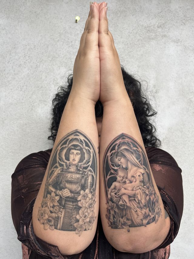 Arms raised together in prayer, each forearm tattooed with a gothic arch — an armoured saint on one, a Madonna with child on the other.