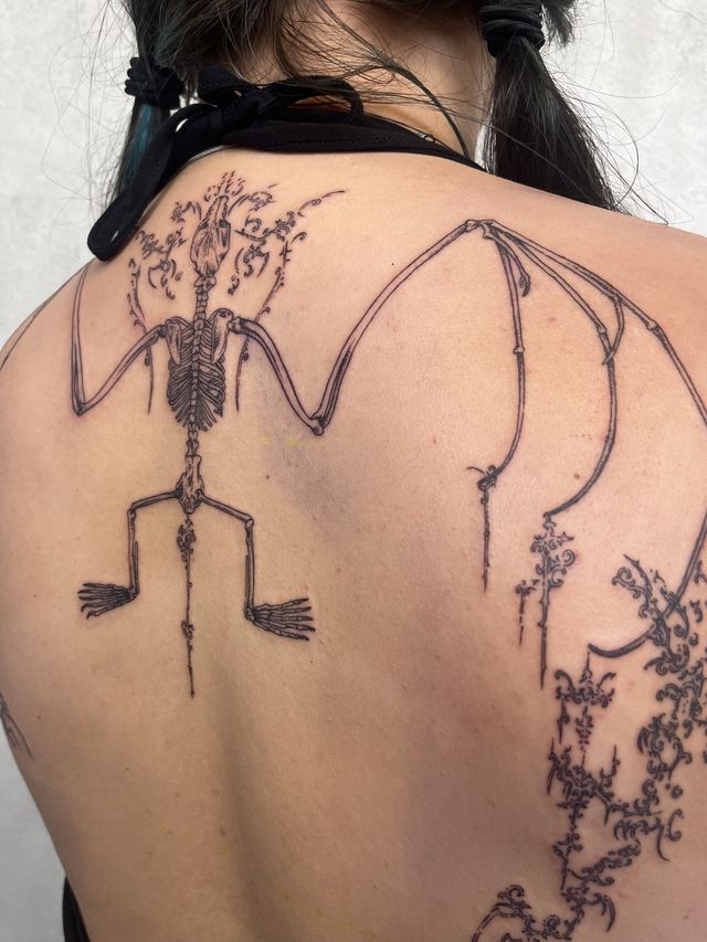 Fine line bat skeleton across the upper back, its wings and spine traced with flowering vines.