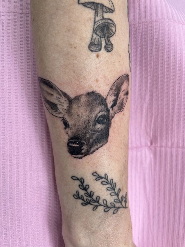 Black and grey deer head with mushrooms above and a leafy sprig below, on an arm.