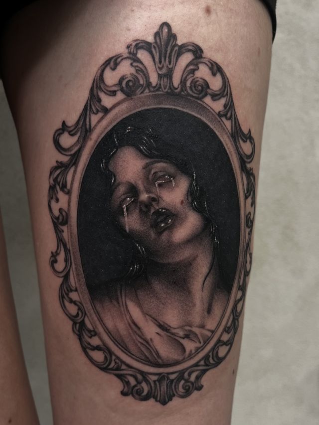 A woman's face inside an ornate oval mirror frame, black and grey on an upper arm.