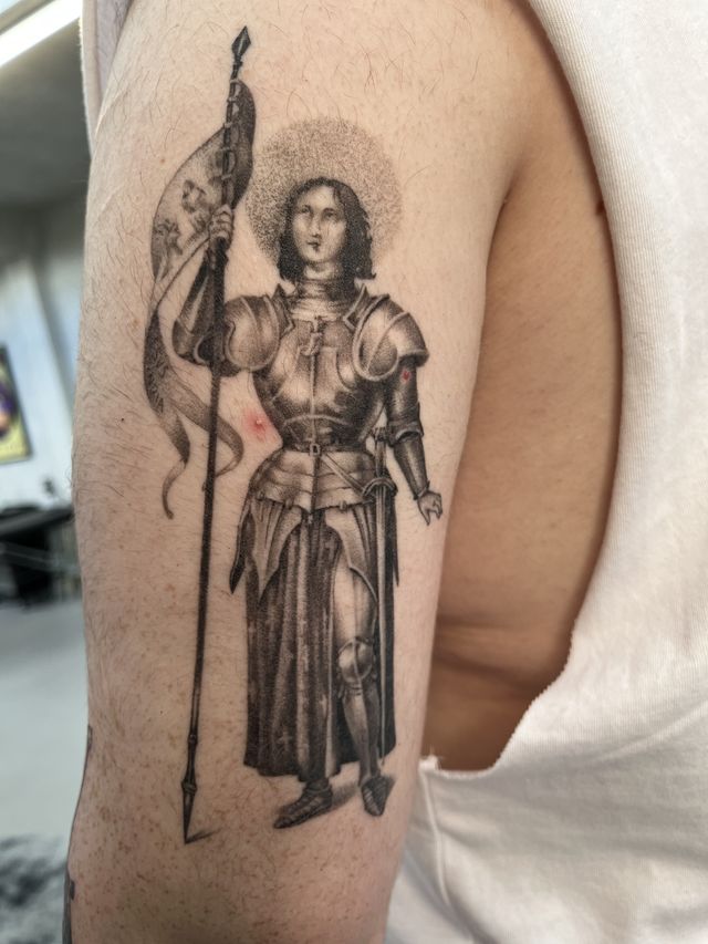 Black and grey armoured Joan of Arc holding a banner and lance, on an upper arm.
