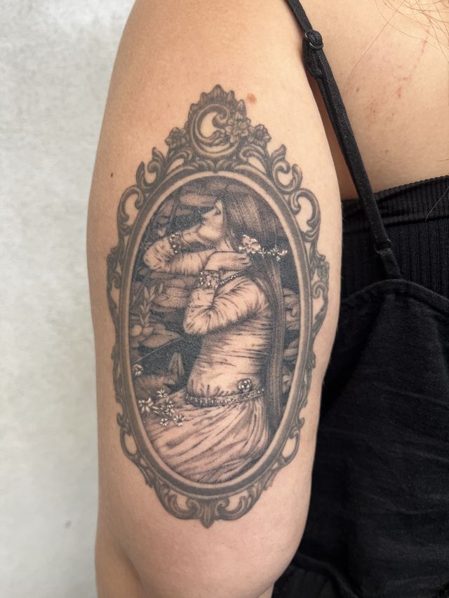 A maiden with long hair inside an ornate Victorian mirror frame, in black and grey on an upper arm.