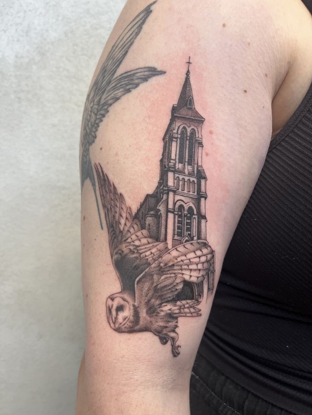 Black and grey barn owl in flight in front of a church spire, on an upper arm.